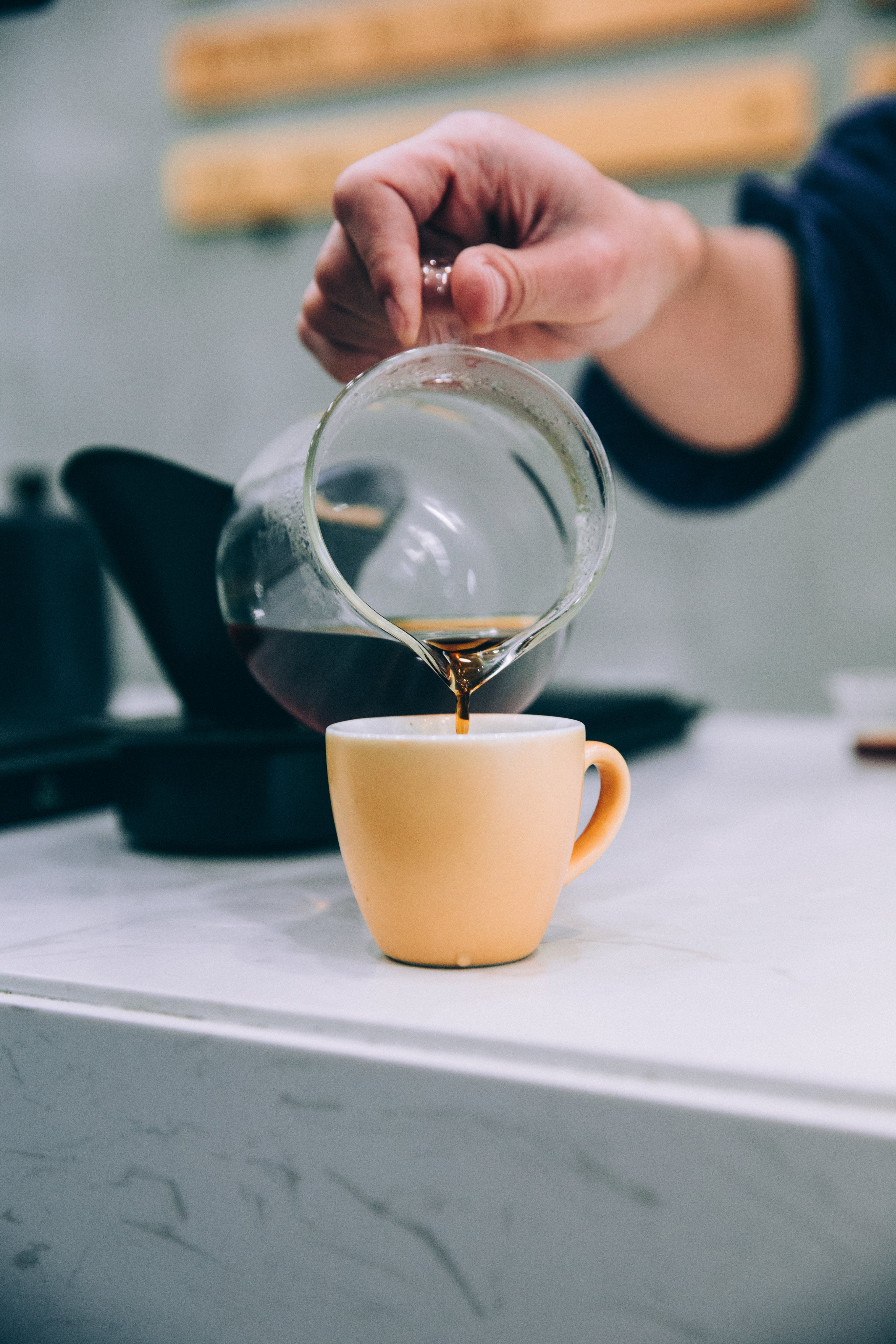 A hand pouring fresh coffee from a glass carafe into a yellow porcelain cup on a white countertop.